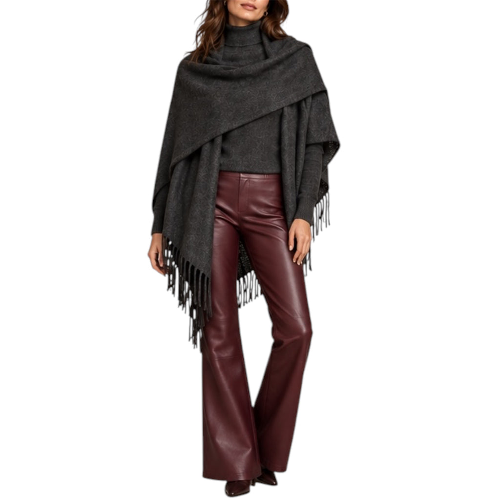 A woman modeling a dark gray knit wrap poncho with fringe, worn over a dark top and paired with dark burgundy faux-leather flare pants, against a plain white background.