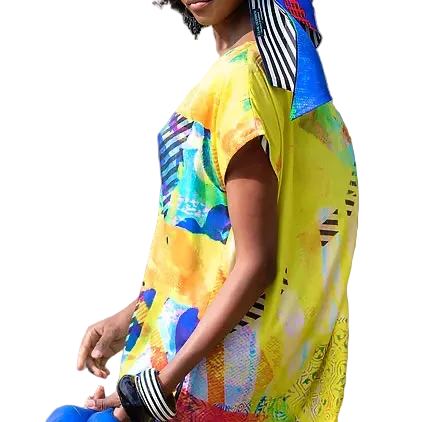A woman wearing a bright yellow abstract-print silk tunic with blue, green, and orange accents, paired with a matching multicolor headscarf, standing outdoors against a blurred sandy background.