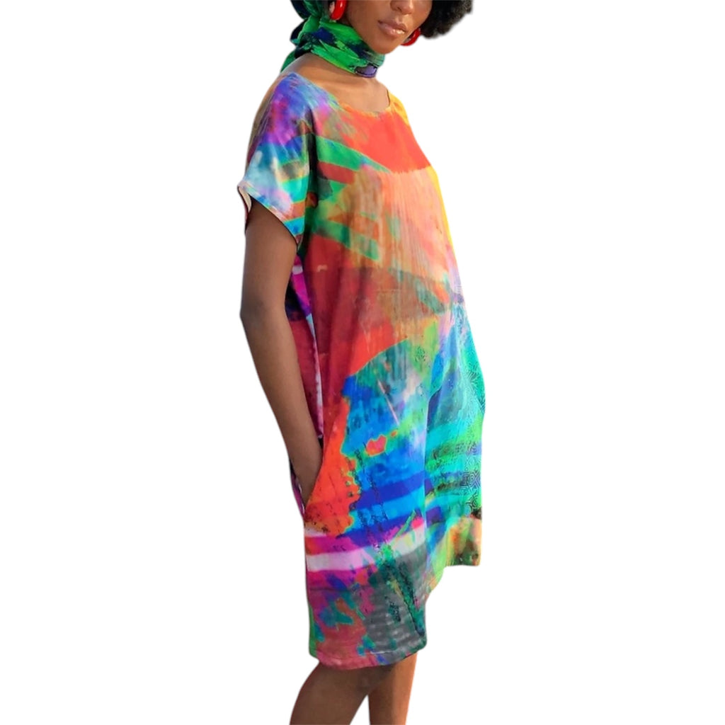 A woman wearing a loose, short-sleeve silk dress by Irish artist Clare O’Connor with an abstract multicolor print in shades of blue, green, orange, pink, and purple, shown against a plain white background.