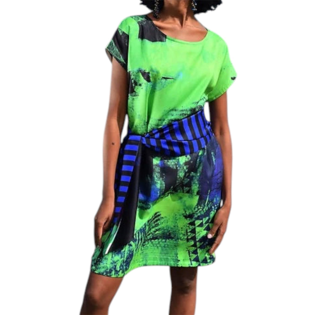 A woman wearing a short-sleeve silk dress by Irish artist Clare O’Connor with a bright green base, black abstract print, and a blue-and-black striped sash tied at the waist, shown against a plain white background.